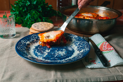 Laddle pouring shakshuka on an empty plate, cooking pan, fresh parsley and gluten free bread