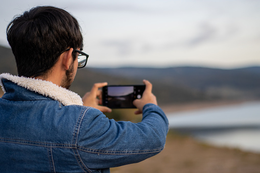 portrait of a young bearded man with glasses taking a picture of the landscape with his cell phone
