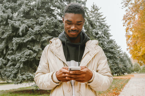 istock Smiling young black man is typing message on smartphone over autumn trees background. 1450439411
