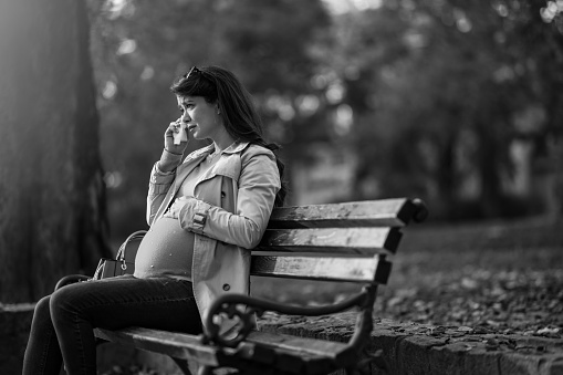 Lonely young pregnant woman crying while sitting in the park on autumn day. She is having problems and have to deal with them alone. Black and white photo.