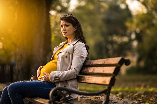 Sad and lonely young pregnant woman sitting in the park and contemplating on autumn day.