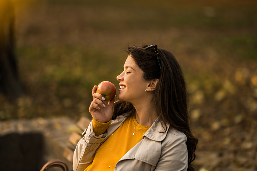 Happy young woman eating an apple while relaxing in the park on autumn day. Young woman makes her health a priority.