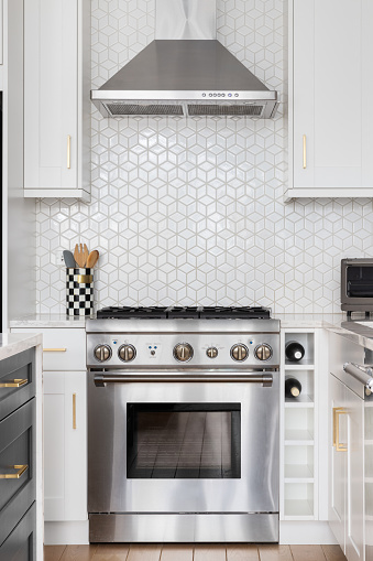 A kitchen stove detail shot in a luxury kitchen with white cabinets, stainless steel appliances, and pattern tile backsplash.