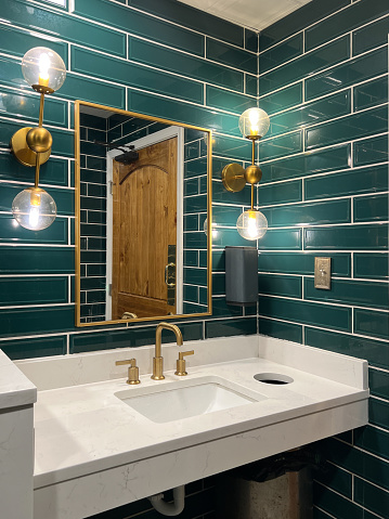 A bathroom with green subway tile walls, gold lights, marble sink and a gold faucet.
