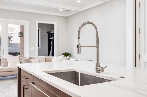 A kitchen sink with water running in a luxurious home looking out towards a living room area. The kitchen island features a white granite and stainless steel tub.