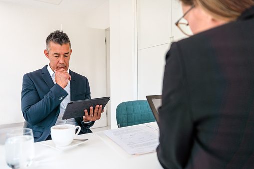 Thoughtful Asian mature man looking at tablet screen. He is seated across female coworker. They both think and talk about the best ideas for the upcoming meeting.