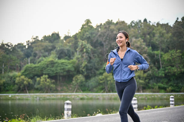 mujer asiática feliz escuchando música mientras disfruta corriendo en el parque por la mañana. - lake asia young women beautiful people fotografías e imágenes de stock