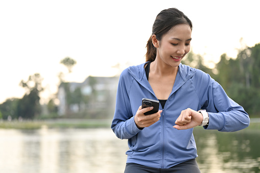 Sporty fit Asian woman in sportswear holding her phone and checking her heart rate on smartwatch.