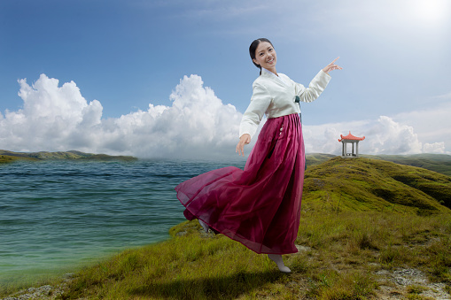 Asian woman wearing a traditional Korean national costume, Hanbok, standing beside the lake with blue sky background