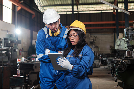 Two industrial workers in safety uniforms and hardhats, male manager, and Black colleague work with tablet to check metalwork machines in manufacturing factory. Professional production engineer team.