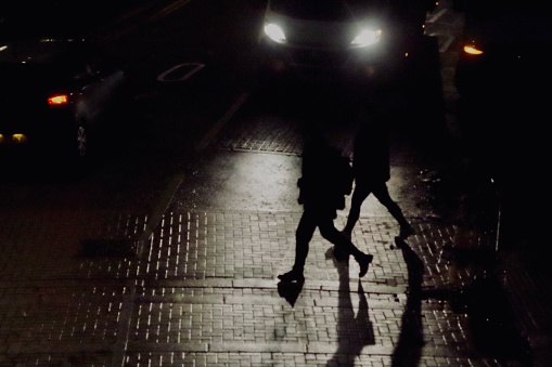 People crossing the road at night lit up by car headlights. Focus on the long shadown cast by the light from the pedestrian