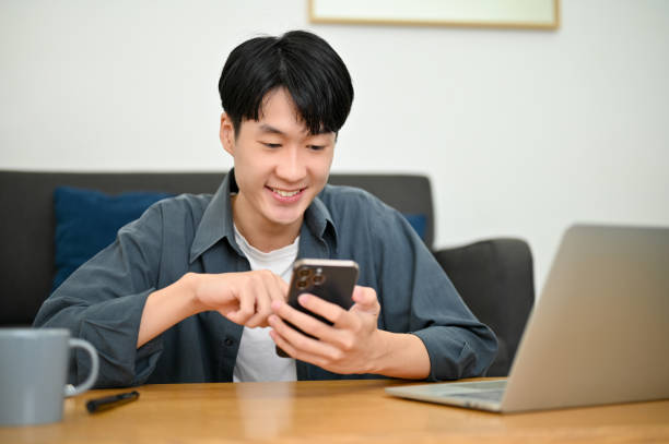 istock Cheerful Asian man using a mobile application on his smartphone while relaxing in the living room. 1450513758