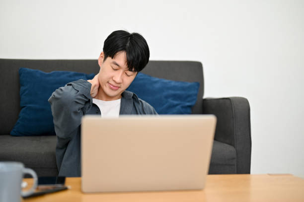 Tired young Asian male freelancer sits in the living room touching his neck, getting neck pain stock photo