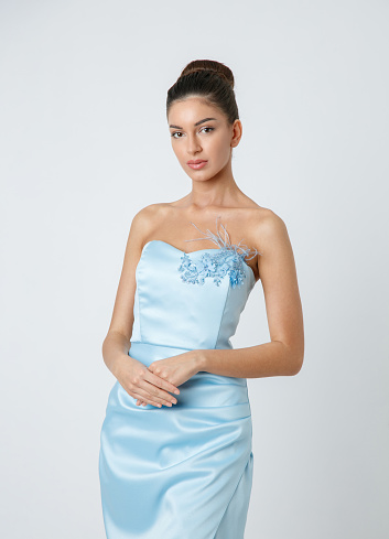 Beautiful young fashion model with brown hair in bun, wearing blue strapless dress looking at camera and holding hand in hand in front, studio shot, beauty and fashion industry