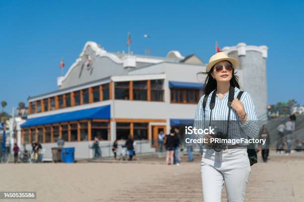 Leisure Stylish Asian Korean Woman Visitor Exploring Santa Monica Beach With Camera On Sunny Day With A Large Local Seafood Restaurant At Background Stock Photo - Download Image Now