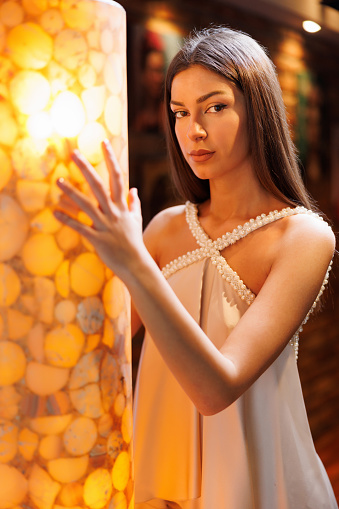 Portrait of beautiful young fashion model in beige sleeveless top holding onto illuminated wall and looking at camera, beauty and fashion industry