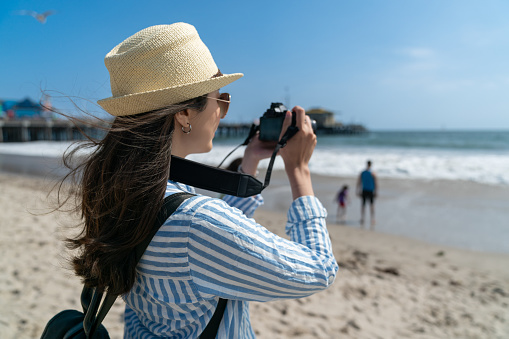 back view of young asian Japanese woman photographer wearing hat taking photos of beautiful ocean waves with slr camera in breeze