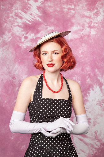 Studio portrait of a cheerful red-haired white woman dressed in retro style in a polka-dot dress against a pink background