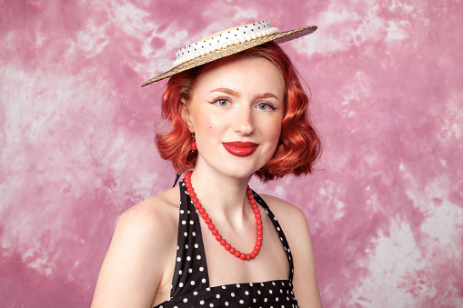 Close up studio portrait of a cheerful red-haired white woman dressed in retro style in a polka-dot dress against a pink background