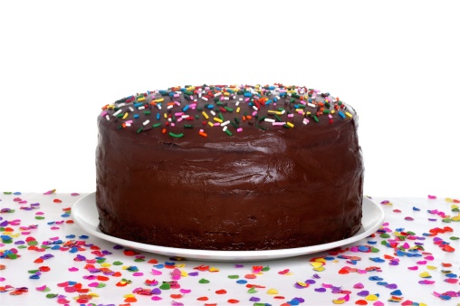 Chocolate cake topped with colorful sprinkles, sitting on a table scattered with confetti.