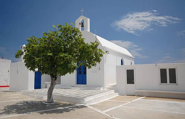 Image shows a church in Serifos, Greece with blue sky and high cloud and tree in foreground