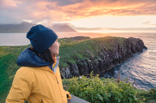 Attractive young asian woman standing and watching puffin bird on the cliff among north arctic ocean in Borgarfjordur Eystri small town on summer at East of Iceland