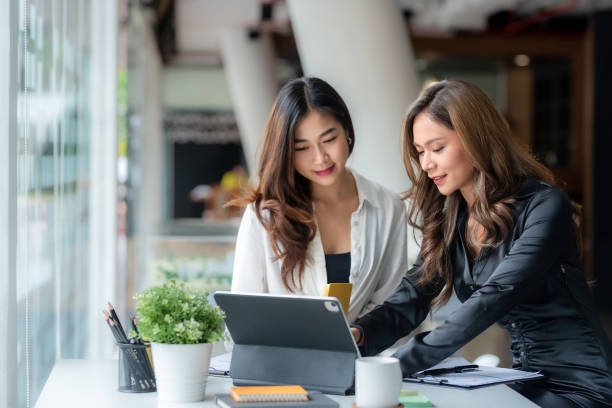 Two beautiful young Asian businesswoman in the conversation, exchanging ideas on laptop computer at work. stock photo