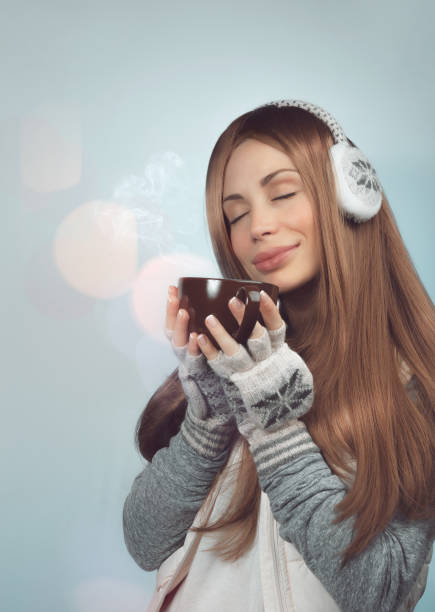 Portrait of a Beautiful Young Woman Enjoying Hot Drink. Nice Female Wearing Warm Earmuffs over Blurry Bokeh Background.