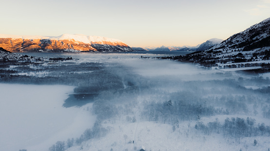 Drone high-angle photo of female in blue jacket contemplating the cold winter morning walking to the wooden hut on the snowy footpath, near the frozen lake and with idyllic view of the snowcapped fjord in Scandinavia