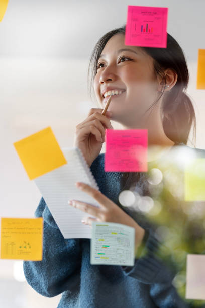 istock Young smiley attractive, businesswoman using post it notes in glass wall to writing strategy business plan to development grow to success 1450623349