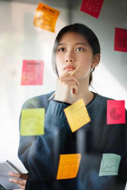 Young businesswoman thinking and planning business strategy using using post it notes on glass wall to development grow to success.