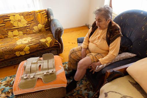 A shot of a senior woman in bad condition, sitting at home and preparing her left leg to wear a leg brace.
