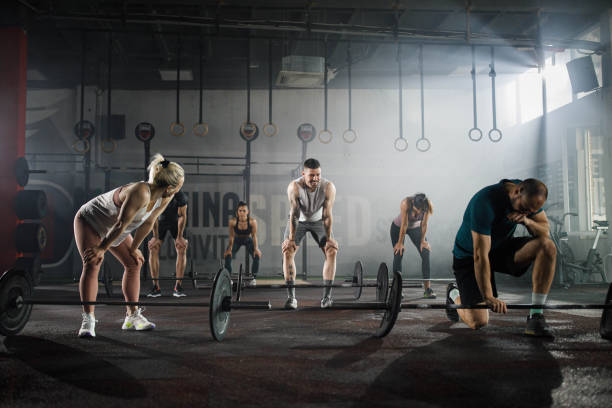 Group of tired athletic people taking a break after exercising with barbells during cross training in a gym. Copy space.