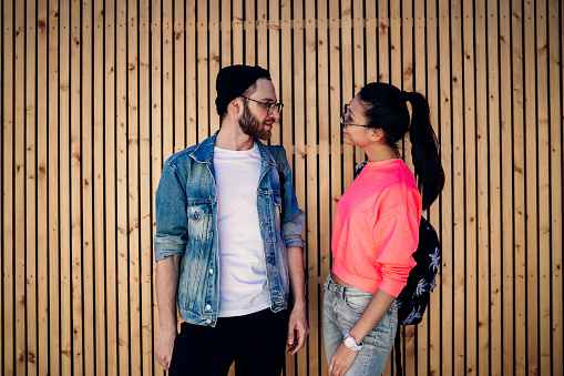 Male and female friends dressed in fashionable streetwear standing near promotional background with copy space for brandname and communicating, trendy hipster guys talking at wooden publicity area