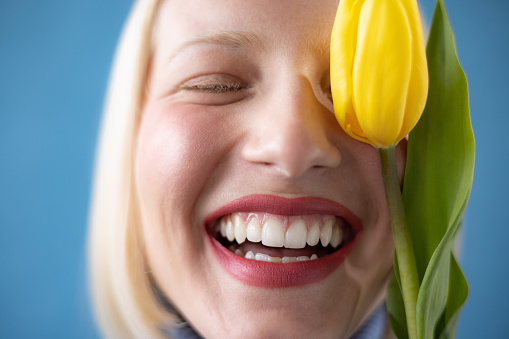 Close up of happy woman with toothy smile hiding her eye with yellow tulip.