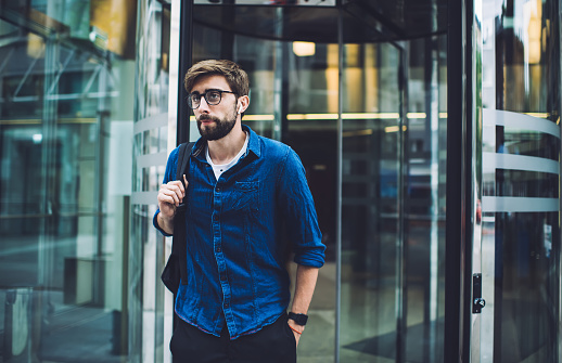 Young bearded male in eyeglasses and casual clothes with backpack over shoulder coming out from office center after working day