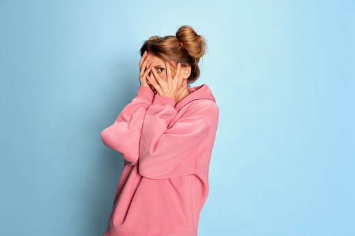 Portrait of young girl in pink hoodie posing, covering face in fear over blue studio background. Emotional stress. Concept of youth, beauty, fashion, lifestyle, emotions, facial expression. Ad