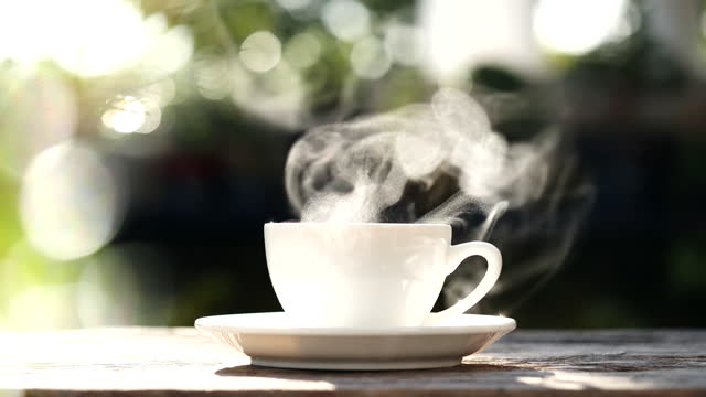 Pouring hot black coffee to white coffee cup, mug with steaming smoke of coffee on old wooden table in morning nature outdoor, garden background, 4K. Hot Drink, Beverage Concept.
