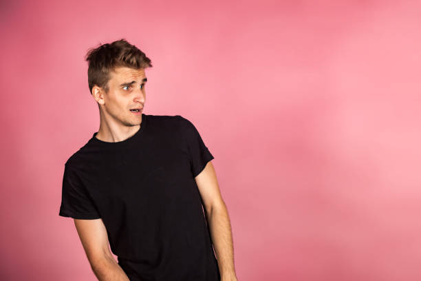 istock Dissatisfied young man looking unpleasantly away, expresses aversion, smirking at pink background. Guy in black t-shirt scared, negative face expressions concept. Advertising. Copy text space 1450678319