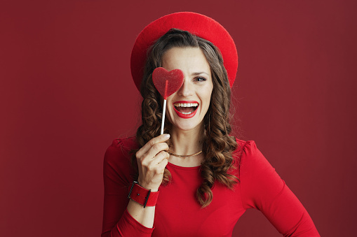 Happy Valentine. happy stylish middle aged woman with long wavy hair in red dress and beret with heart shaped candy on stick isolated on red.