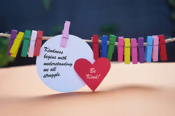 Life inspirational motivational quote - Kindness begins with understanding we all struggle. Be kind. Text message on red heart and white circle paper with colorful wooden clips on orange background.