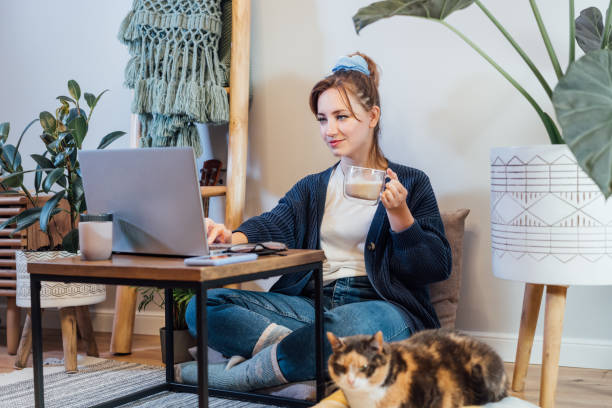 istock Woman using laptop while sitting on floor cushions with sleeping cat pet. Cozy workspace in modern Scandi interior home with green plants. Remote work at home. Online education or entertainment 1450690875