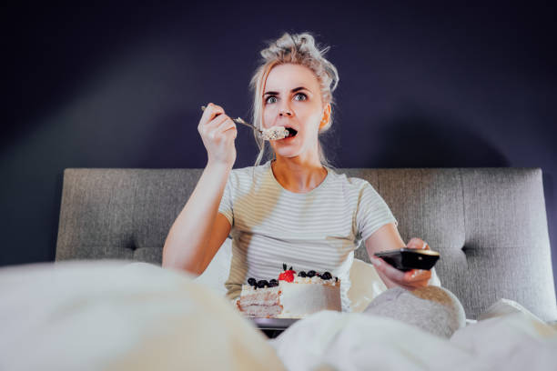 istock Shocked, stressed, surprised woman eating cake and holding a remote control while watching TV in bed. Scary horror movie or terrible news, scare in her eyes Overeating, Jamming of negative emotions 1450690900