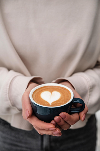 Girl in a biege pullover holds a cup of cappuccino with heart on top. Sant Valentine Day concept
