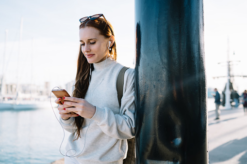 Attractive smiling thoughtful adult female in headphones and sunglasses focusing on screen and interacting with smartphone while standing and leaning on lamp post during sunny day on blurred background of ships in harbor
