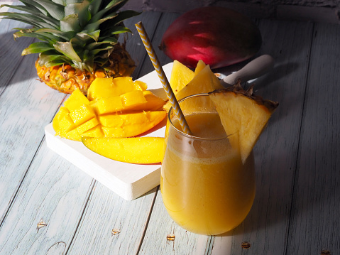 Glass of fresh mango and pineapple juice, slices of mango and pineapple fruit on a wooden background. Vertical image