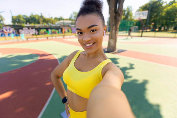 Smiling African American woman taking selfie using cell phone gadget, millennial sports influencer recording video blog. Positive black lady holding camera taking break standing outside at city field