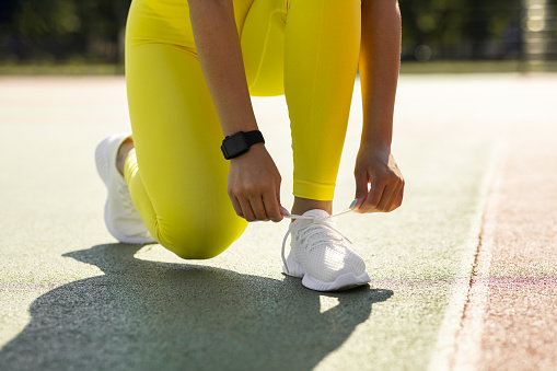 Training Concept. Closeup cropped view of unrecognizable black female athlete in bright yellow leggings tying her shoelaces before jogging outdoors in the morning, wearing white sneakers