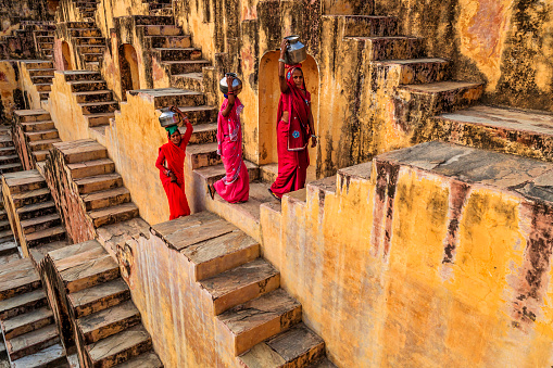 Indian women carrying water from stepwell near Jaipur, Rajasthan, India. Women and children often walk long distances to bring back jugs of water that they carry on their head. 
Stepwells are wells in which the water may be reached by descending a set of steps.