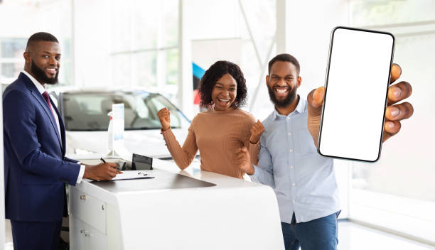 自動車保険。自動車ショールームに立ち、空白のスマートフォンを見せる黒人の配偶者 - car african descent shopping car dealership ストックフォトと画像
